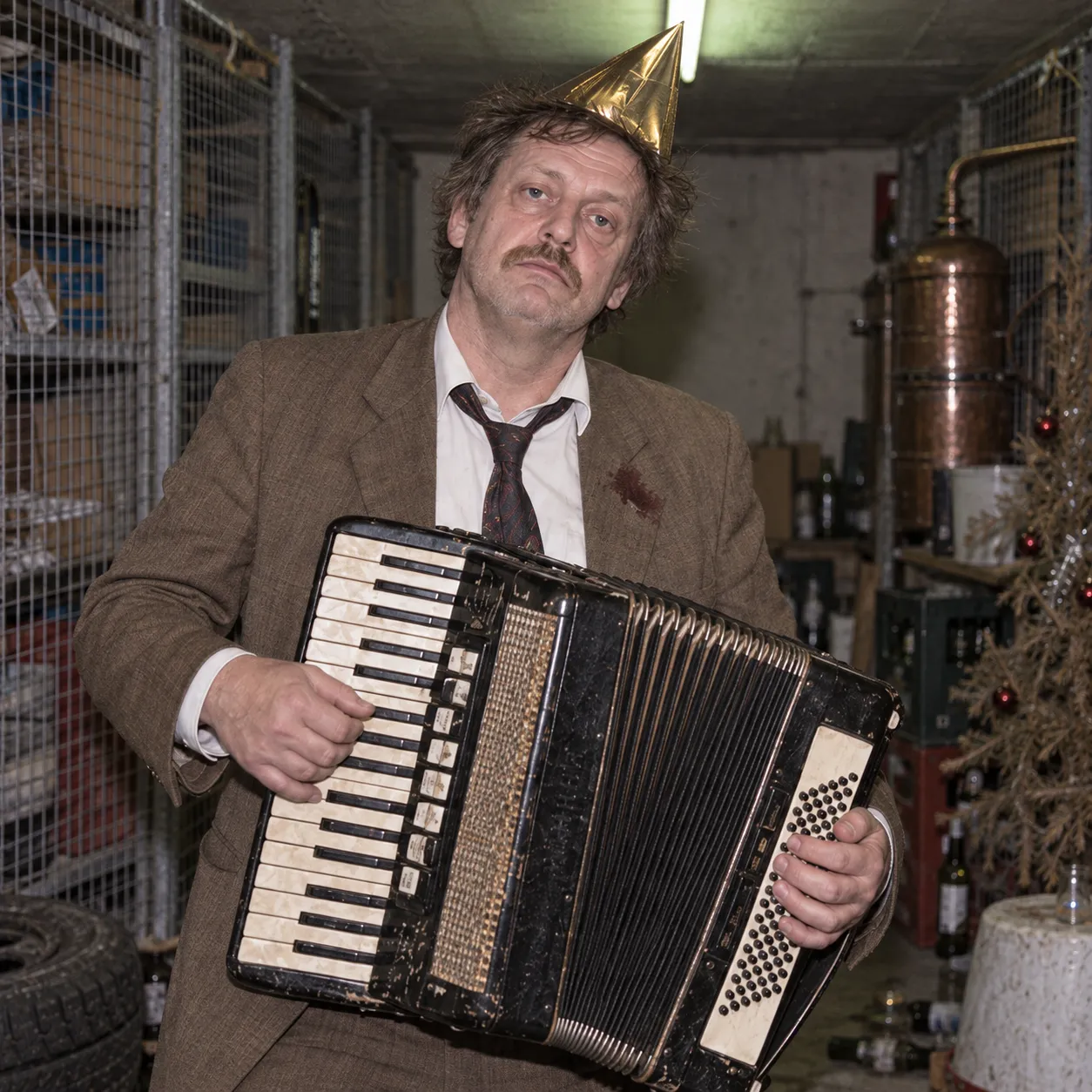 Bängt Ljungqvist in a stained brown jacket and gold party hat, playing a battered accordion